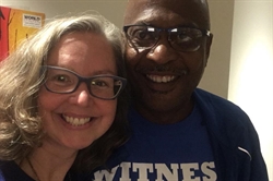 [ai] A woman with glasses smiling next to a man wearing glasses and a blue shirt, both looking at the camera. The background features colorful artwork.