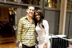 [ai] Two individuals posing for a photograph at an event. One person is wearing a patterned yellow shirt, and the other is in a fitted light-colored dress. The background shows a gathering area with tables.