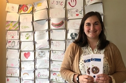 [ai] A woman holding brochures poses in front of a wall filled with colorful drawings and messages promoting equality and education.