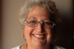 [ai] A smiling woman with curly gray hair wearing glasses and silver earrings, posed against a neutral background. She is dressed in a light top and appears to be in a well-lit setting.