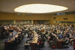 [ai] A large audience gathered in a formal conference room, seated in rows with desks. The room is well-lit, featuring a circular ceiling and wooden paneling, indicating a significant meeting or event.