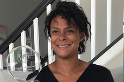 [ai] A woman with curly hair smiling while holding a crystal award in a well-lit room with white walls and a staircase in the background.