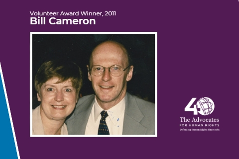 [ai] A man and a woman smiling together in a formal portrait. The man is wearing glasses and a suit, while the woman has short hair and is wearing a light-colored blouse. Background includes a design for an award and text indicating "Volunteer Award Winner, 2011" and the name "Bill Cameron."