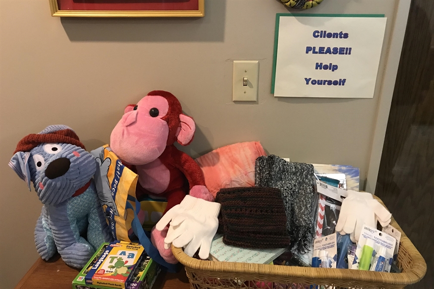 [ai] A display of a blue stuffed dog and a pink stuffed monkey beside a basket filled with hats, gloves, and various small items, with a sign stating 'Clients PLEASE! Help Yourself' on the wall.