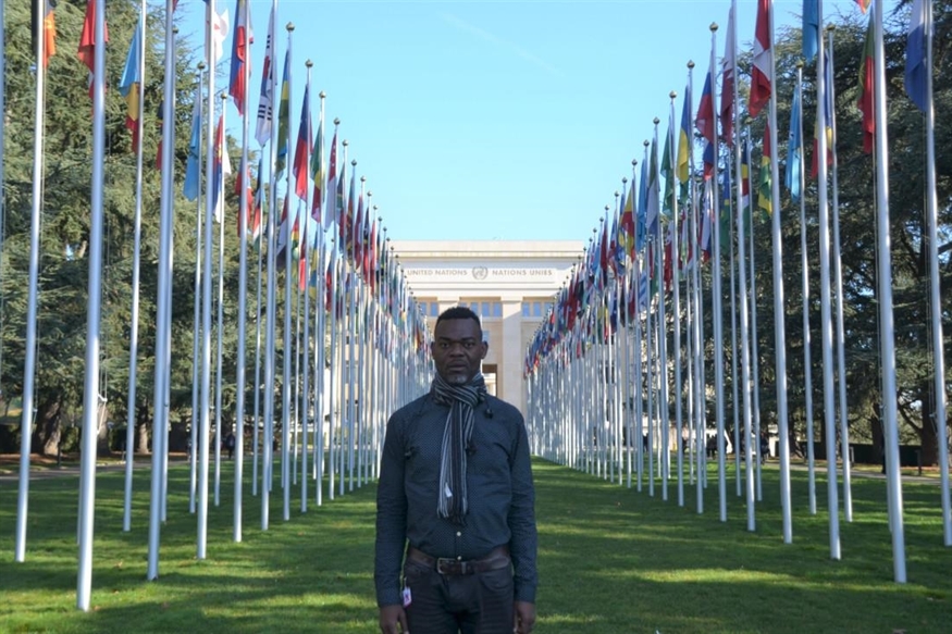 [ai] A person standing in front of a row of flagpoles displaying various national flags in a park-like setting, with a large building visible in the background.