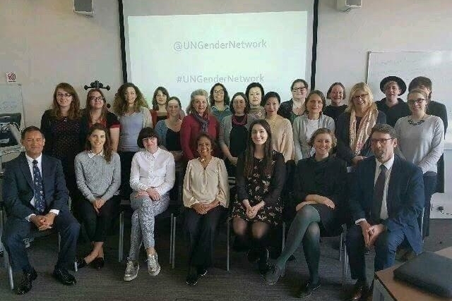 [ai] A diverse group of individuals posing for a photo in a professional setting, with a projection screen in the background displaying the @UNGenderNetwork and #UNGenderNetwork. The group includes men and women, dressed in formal attire.
