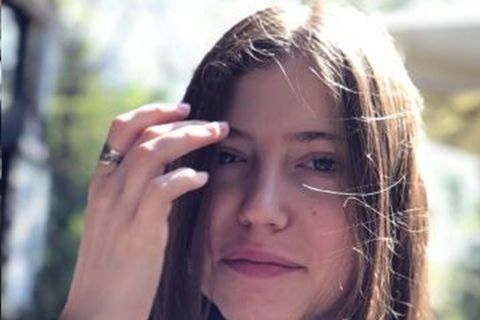 [ai] A young woman with long, straight hair lightly touching her face while sitting outdoors, surrounded by greenery. Sunlight casts a soft glow on her features.