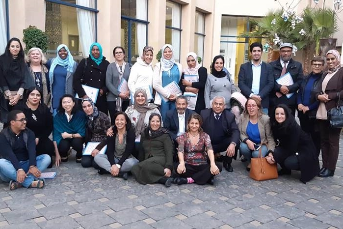 [ai] A diverse group of individuals, both men and women, standing and sitting together outdoors. They are smiling and holding documents, gathered in front of a building with large windows and greenery.