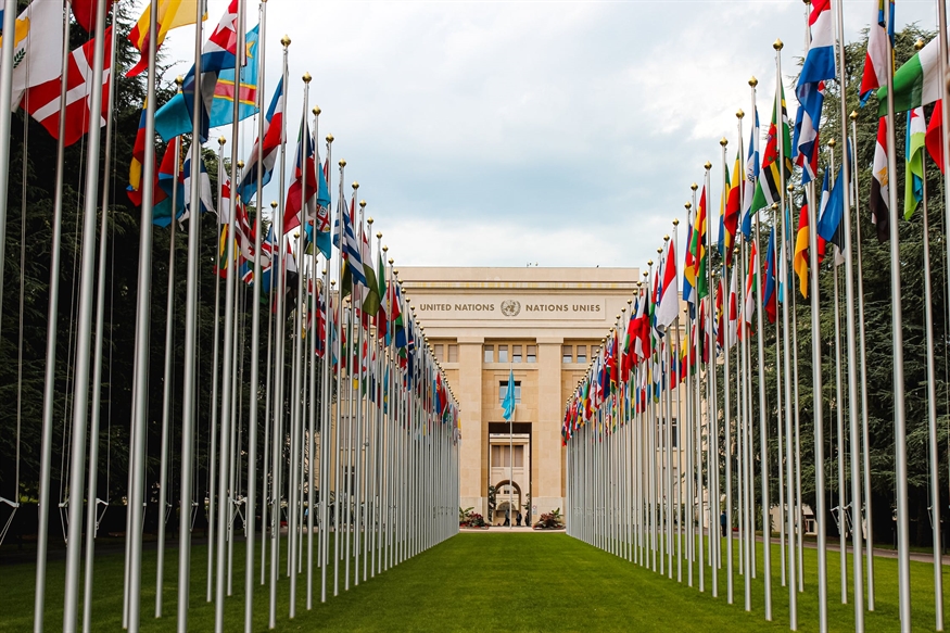 [ai] A view of the United Nations building, framed by rows of international flags on flagpoles, set in a green lawn under a cloudy sky.
