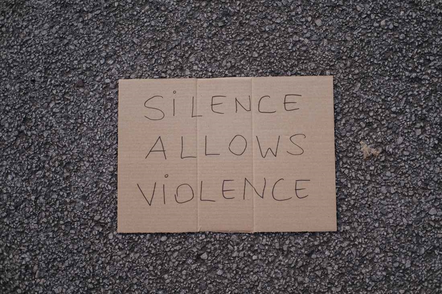 [ai] A cardboard sign with handwritten text reading "SILENCE ALLOWS VIOLENCE" placed on a textured asphalt surface.