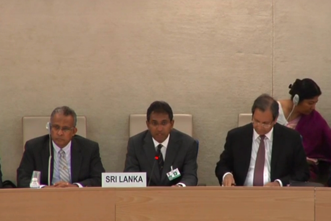 [ai] Three men seated at a conference table, with one man speaking into a microphone. A placard reading 'SRI LANKA' is visible in front of him. In the background, a woman wearing headphones is seated.