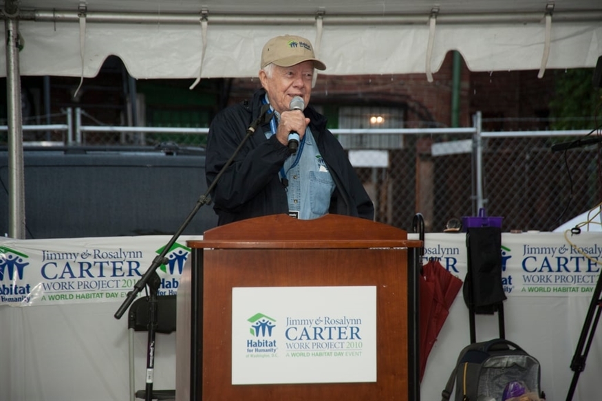 [ai] An older man speaking into a microphone at a podium under a tent. He is wearing a cap and a jacket. Behind him, there are banners for Habitat for Humanity and the Jimmy & Rosalynn Carter Work Project.